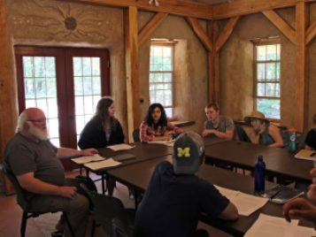 A white-haired professor with a bushy beard sit at a collection of tables where undergraduate students gather for class inside the straw bale building.